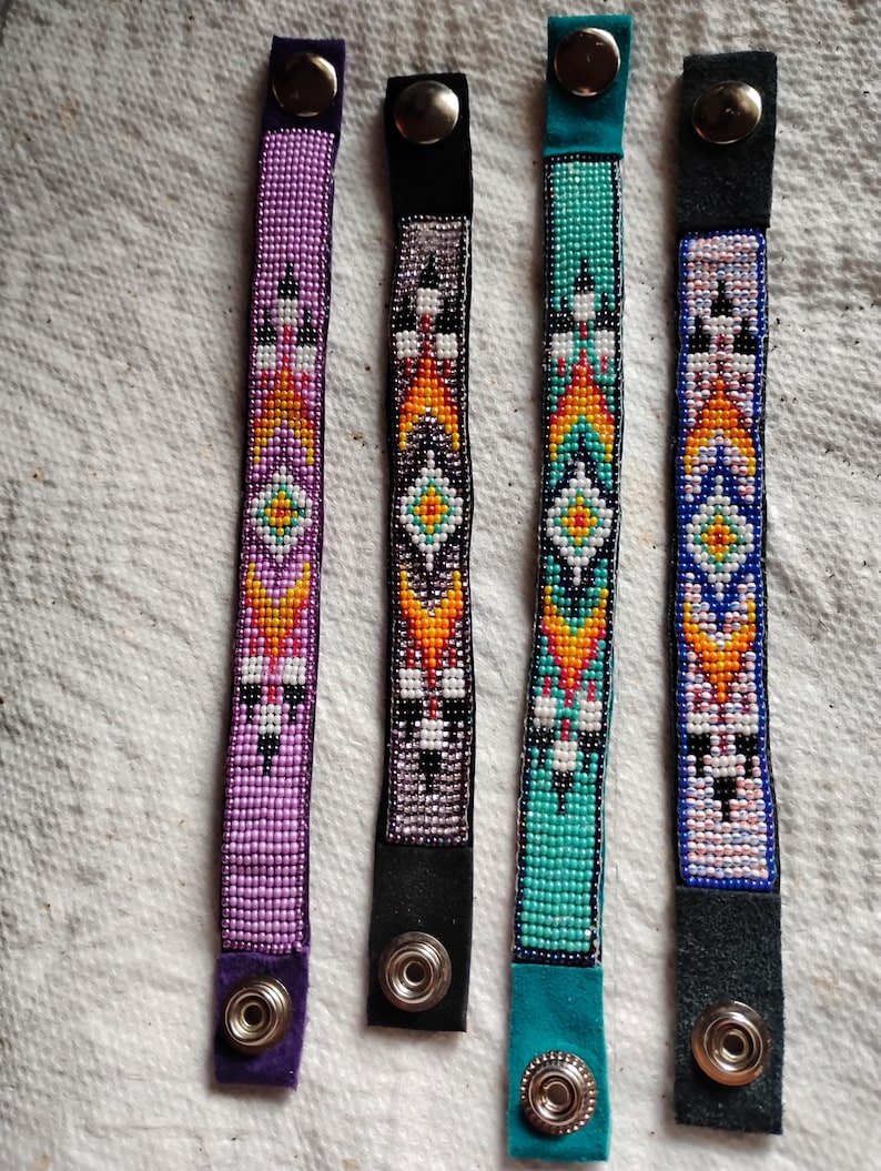K&ouml;nnte beinhalten: Vier Perlenarmb&auml;nder mit bunten geometrischen Mustern und Lederriemen mit Druckkn&ouml;pfen. Die Armb&auml;nder sind in den Farben Lila, T&uuml;rkis, Schwarz und Wei&szlig;.