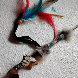 May include: A wooden pipe with an eagle head carving, decorated with beads and feathers. The pipe is brown with a black stem and a gray fur trim. The feathers are red, blue, and white.