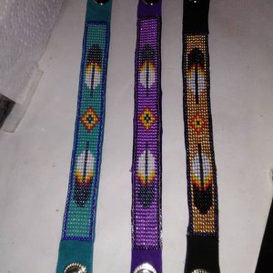 Könnte beinhalten: Drei Perlenarmbänder mit Lederriemen und Druckknöpfen. Die Armbänder zeigen ein Federdesign in Türkis, Lila und Gold.