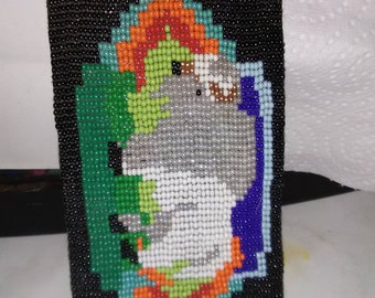 May include: A beaded bracelet with a colorful design featuring a gray animal with a white face. The design is surrounded by green, orange, and blue beads.