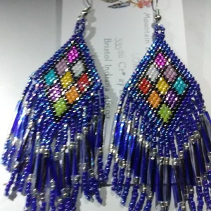 May include: A pair of blue beaded earrings with a colorful geometric pattern and silver fringe. The earrings are handmade and feature a variety of colors, including red, orange, yellow, green, blue, purple, and white.