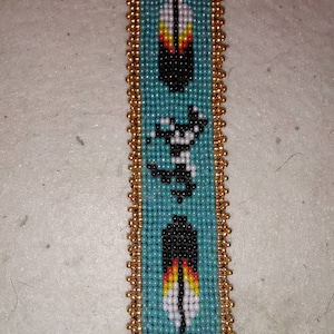 May include: A beaded bookmark with a turquoise background and gold trim. The bookmark features three black and white feathers with red and yellow accents.