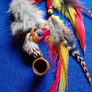 May include: A handcrafted tobacco pipe with an eagle head carving, adorned with gray feathers and colorful red, yellow, and black feathers. The pipe has a wooden bowl and is set against a blue background.
