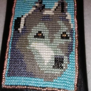 May include: A close-up of a beaded picture of a wolf's face. The wolf is gray and white with brown eyes. The picture is framed in a black frame with a copper-colored border.