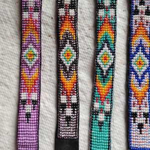 K&ouml;nnte beinhalten: Vier Perlenarmb&auml;nder mit bunten geometrischen Mustern. Die Armb&auml;nder sind aus kleinen Perlen in den Farben Lila, T&uuml;rkis, Schwarz, Wei&szlig;, Orange und Gelb gefertigt. Jedes Armband hat ein schwarzes Lederband mit einem Druckknopfverschluss.