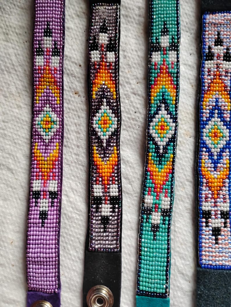 K&ouml;nnte beinhalten: Vier Perlenarmb&auml;nder mit bunten geometrischen Mustern. Die Armb&auml;nder sind aus kleinen Perlen in den Farben Lila, T&uuml;rkis, Schwarz, Wei&szlig;, Orange und Gelb gefertigt. Jedes Armband hat ein schwarzes Lederband mit einem Druckknopfverschluss.