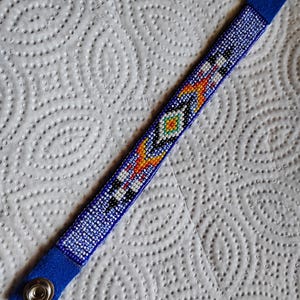 K&ouml;nnte beinhalten: Ein blaues Perlenarmband mit einem farbenfrohen geometrischen Muster. Das Armband hat an jedem Ende einen Druckknopfverschluss.