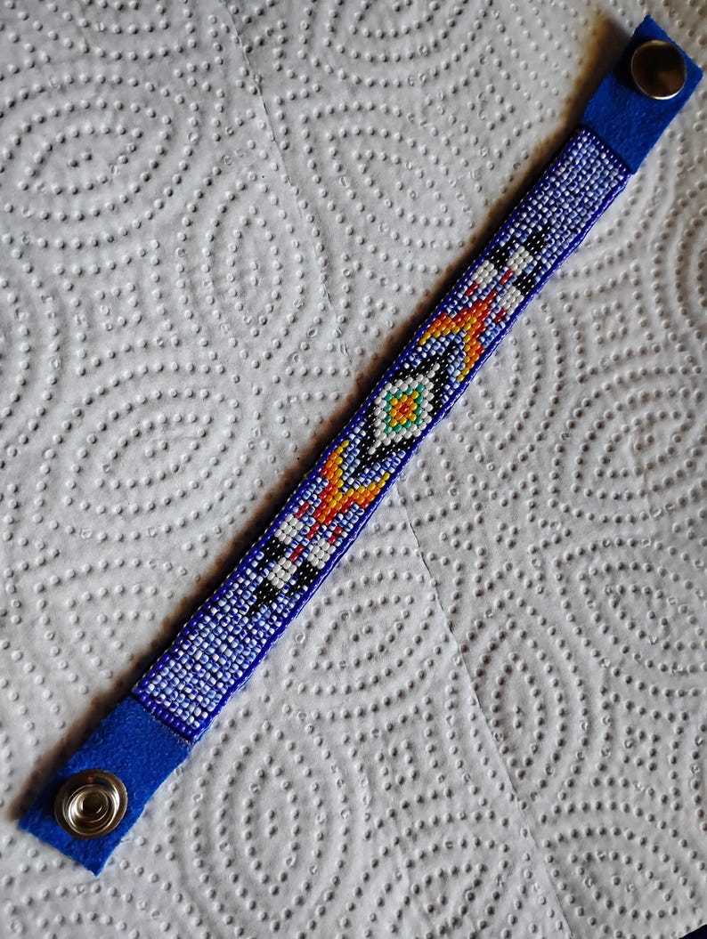 K&ouml;nnte beinhalten: Ein blaues Perlenarmband mit einem farbenfrohen geometrischen Muster. Das Armband hat an jedem Ende einen Druckknopfverschluss.