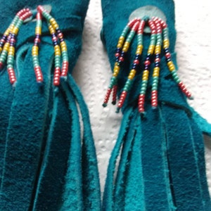 May include: Two teal blue leather tassels with fringe and colorful beaded accents. The tassels are tied together with a leather cord.