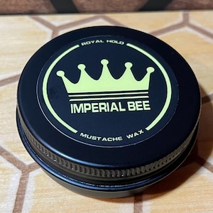 May include: A round black metal tin of Imperial Bee mustache wax with a screw-top lid. The lid features a circular label with a light yellow crown graphic, the text Royal Hold, Imperial Bee, and Mustache Wax printed in a bold font.