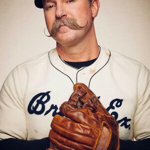 May include: A vintage-style baseball uniform featuring a cream-colored jersey with dark blue script text reading Br Ex, a black undershirt, and a white cap with a black brim. A worn brown leather baseball glove is held in front of the chest.