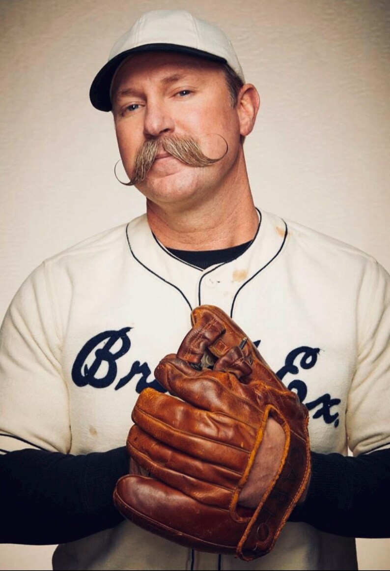 May include: A vintage-style baseball uniform featuring a cream-colored jersey with dark blue script text reading Br Ex, a black undershirt, and a white cap with a black brim. A worn brown leather baseball glove is held in front of the chest.