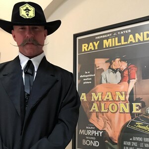 May include: A black cowboy hat featuring a yellow hexagonal patch with a bee icon and the text Imperial Bee, worn with a black suit jacket, white collared shirt, and a black bolo tie, standing beside a framed movie poster for A Man Alone.