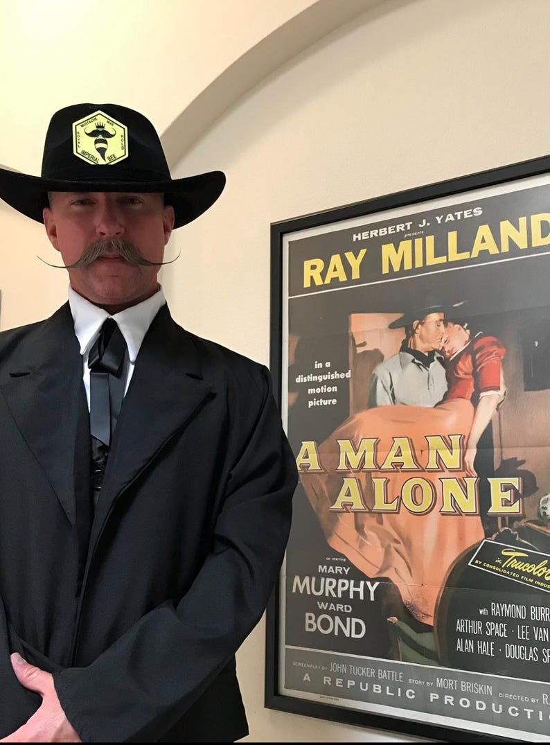 May include: A black cowboy hat featuring a yellow hexagonal patch with a bee icon and the text Imperial Bee, worn with a black suit jacket, white collared shirt, and a black bolo tie, standing beside a framed movie poster for A Man Alone.