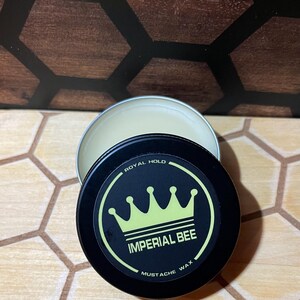 May include: A round black metal tin containing mustache wax with a lid featuring a yellow crown graphic and the text Imperial Bee Royal Hold Mustache Wax. The tin is partially open, revealing the light-colored wax inside the container.