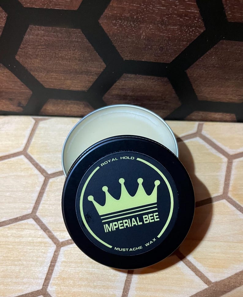 May include: A round black metal tin containing mustache wax with a lid featuring a yellow crown graphic and the text Imperial Bee Royal Hold Mustache Wax. The tin is partially open, revealing the light-colored wax inside the container.