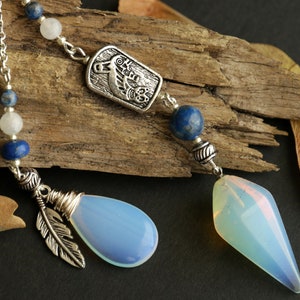 Puede incluir: Dos péndulos de cristal con cadenas y detalles plateados. Uno tiene un cristal azul claro en forma de lágrima con un dije de pluma plateada. El otro tiene un cristal azul claro facetado. Ambos tienen cuentas azules y blancas.