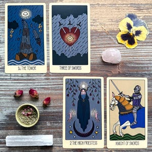 a collection of tarot cards and various objects arranged on a wooden surface. The cards feature illustrations of a tower, three swords, a knight on horseback, and a high priestess. There are also rose petals, a crystal, and a flower in the scene.
