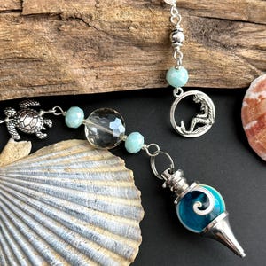 May include: A silver and blue crystal pendulum with a mermaid charm, a turtle charm, and clear and aqua blue beads. The pendulum is hanging from a silver chain.