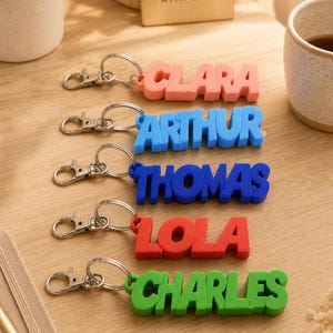 Peut inclure: Porte-clés personnalisés avec des noms colorés : "Clara" rose, "Arthur" bleu, "Thomas" bleu, "Lola" rouge et "Charles" vert. Chaque porte-clés a un fermoir et un anneau argentés. Une pancarte en bois dit "HANDMADE WITH LOVE".
