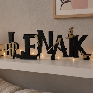 May include: Black wooden letters spelling "LENAK" with decorative accents, including a lightning bolt and a wizard's hat. The letters sit on a white shelf with fairy lights, books, a succulent, and a pink pillow.