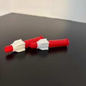 Könnte beinhalten: Zwei rote Kunststoff-Applikatorspitzen mit weißen Kunststoffkappen. Die Spitzen sind zum Abgeben von Flüssigkeiten oder Cremes konzipiert.