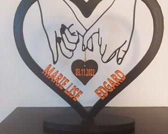 May include: Black metal heart-shaped stand with two hands holding each other's pinky fingers. The stand has a small black heart hanging from it with the date "03.11.2022" and the names "Marie Lise" and "Edgard" written on the stand.