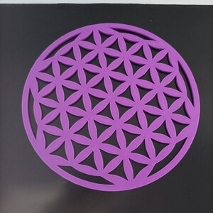 Op de afbeelding: Een rond, paars, decoratief object met een geometrisch patroon van overlappende cirkels en driehoeken. Het patroon staat bekend als de Bloem des Levens.