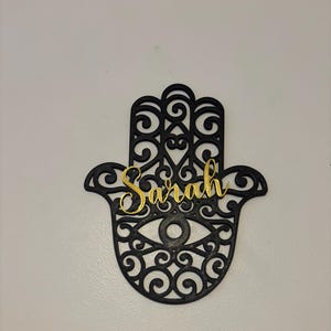 Può includere: Taglio a mano di hamsa nero con dettagli intricati e una targhetta con il nome "Sarah" dorata.