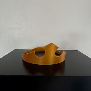 Può includere: Una corona di plastica dorata con un design semplice. La corona è su una superficie nera.