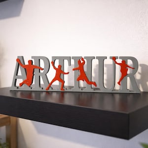 Peut inclure: Un panneau en bois gris épelant "ARTHUR" avec des silhouettes orange de joueurs de basket-ball. Le panneau est affiché sur une étagère noire contre un mur blanc. Article de décoration pour une chambre d'enfant ou un thème sportif.