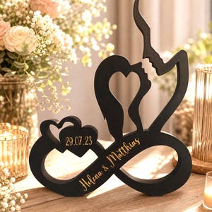 Può includere: Scultura in legno nero raffigurante due profili che si fronteggiano, formando un cuore. Include un simbolo dell'infinito, due cuori con la data 29.07.23 e i nomi "Helena & Matthias" in lettere dorate. Posizionata su una superficie di legno.
