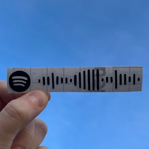 May include: A white paper square with a black Spotify logo and a black and white barcode. The square is being held in a hand against a blue sky.