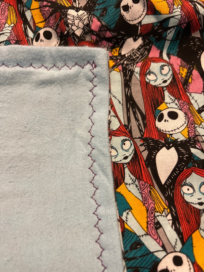 Peut inclure: Un tissu bleu clair avec une bordure cousue en zigzag violet est &agrave; c&ocirc;t&eacute; d'un tissu &agrave; motifs repr&eacute;sentant des personnages de L'&Eacute;trange No&euml;l de Monsieur Jack. Le tissu &agrave; motifs a un fond noir avec des images de Jack Skellington et Sally.