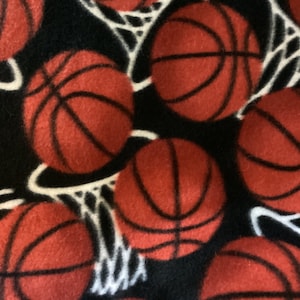 May include: A black fleece fabric featuring a repeating pattern of red basketballs and white basketball nets. The basketballs have black lines. The bottom edge has a decorative black trim with red stitching. This fabric is suitable for sports-themed projects.