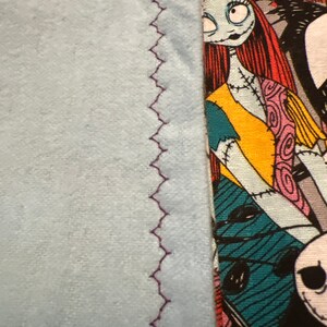 Peut inclure: Gros plan sur un article en tissu. Le c&ocirc;t&eacute; gauche pr&eacute;sente un mat&eacute;riau bleu clair avec une couture en zigzag violet. Le c&ocirc;t&eacute; droit affiche un tissu &agrave; motifs avec un personnage de L'&Eacute;trange No&euml;l de Monsieur Jack, avec un visage bleu clair, des cheveux roux et un motif patchwork.