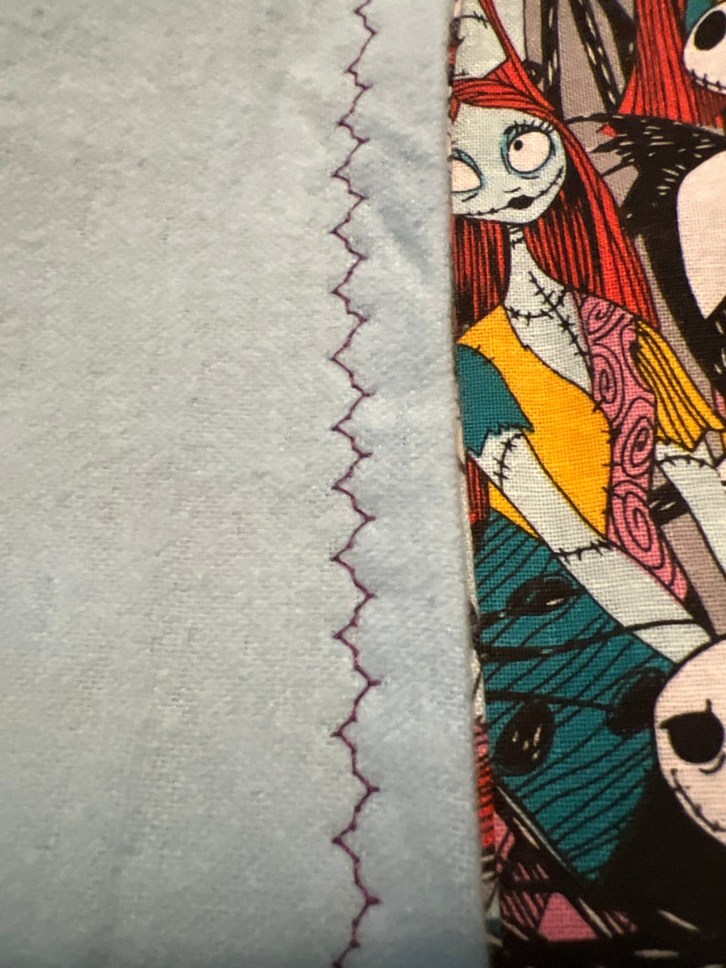 Peut inclure: Gros plan sur un article en tissu. Le c&ocirc;t&eacute; gauche pr&eacute;sente un mat&eacute;riau bleu clair avec une couture en zigzag violet. Le c&ocirc;t&eacute; droit affiche un tissu &agrave; motifs avec un personnage de L'&Eacute;trange No&euml;l de Monsieur Jack, avec un visage bleu clair, des cheveux roux et un motif patchwork.