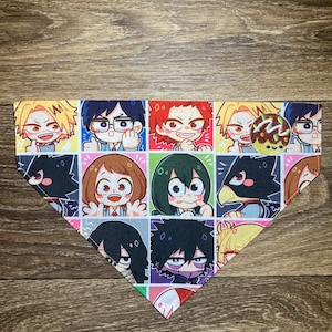 May include: A colorful pet bandana featuring a repeating pattern of anime-style character portraits in a grid. The characters have various hair colors and expressions, set against a white background with colorful squares. The bandana is triangular.