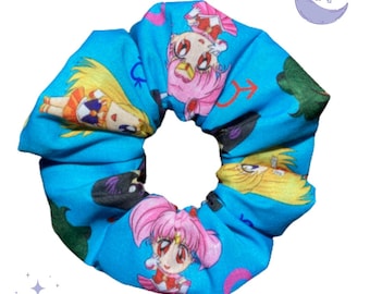 Op de afbeelding: Een blauwe scrunchie met een herhaald patroon van anime-personages. De personages hebben roze, gele en zwarte haren en staan tegen een blauwe en groene achtergrond. Een maan en sterren staan op de achtergrond.