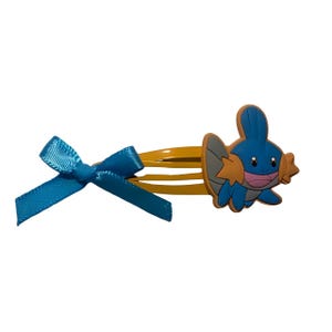 May include: A yellow metal hair clip featuring a blue ribbon bow on one end and a blue and orange cartoon creature character on the other. The character has a blue body, orange fins, and a pink mouth, attached to the side of the hair clip.