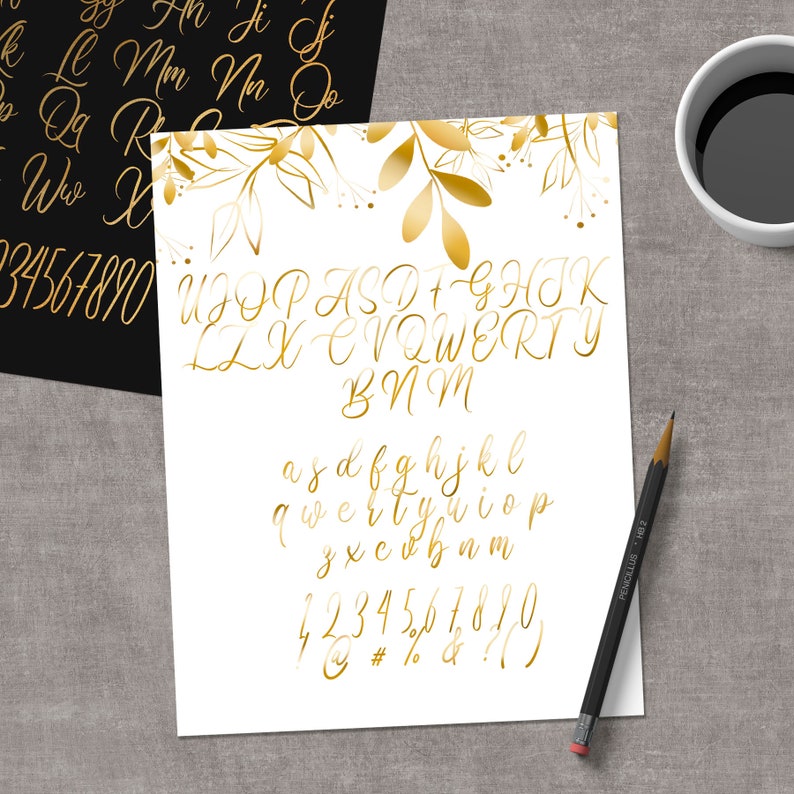 May include: A white sheet of paper with gold calligraphy of the alphabet, numbers, and punctuation marks. The letters are in a cursive font and are arranged in rows. The paper is decorated with gold leaf branches and leaves.