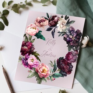 May include: A wedding invitation with a floral wreath design. The wreath is made of pink, purple, and green flowers. The text "Kelly & Andrew" is in the center of the wreath.