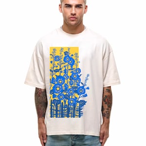 May include: A white t-shirt with a blue and yellow floral print. The print features a fence and flowers in a yellow background.