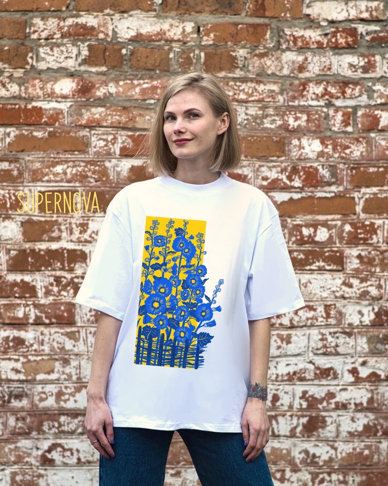 May include: White t-shirt with a graphic print of blue flowers in a yellow rectangle. The text "SUPERNOVA" is visible in the top left corner of the image.