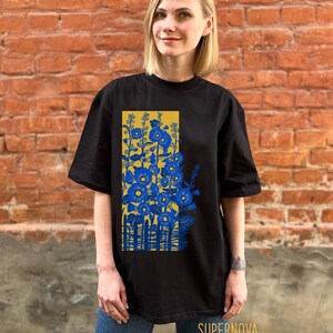 May include: Black t-shirt with a graphic print of blue flowers and a yellow fence. The text "SUPERNOVA" is printed on the bottom of the shirt.