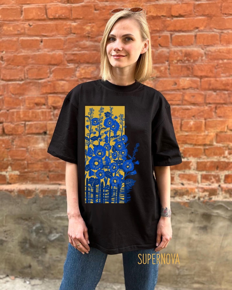 May include: Black t-shirt with a graphic print of blue flowers and a yellow fence. The text "SUPERNOVA" is printed on the bottom of the shirt.
