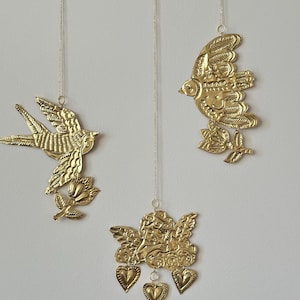 Puede incluir: Tres adornos de metal dorado colgando de cuerdas. Los adornos tienen forma de pájaros y un querubín con corazones. El adorno del querubín tiene una perla colgando de la parte inferior.