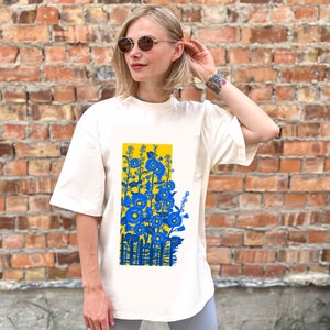 May include: A white t-shirt with a blue and yellow floral print. The print features a fence and flowers in a yellow field.