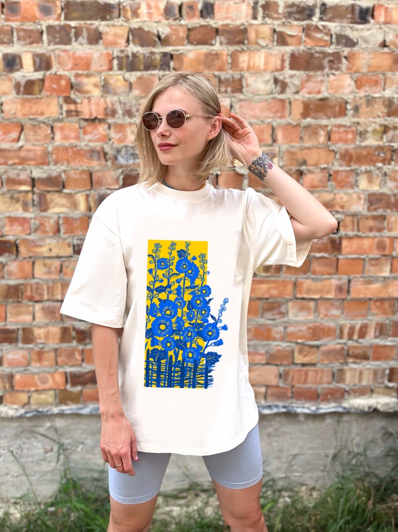 May include: A white t-shirt with a blue and yellow floral print. The print features a fence and flowers in a yellow field.