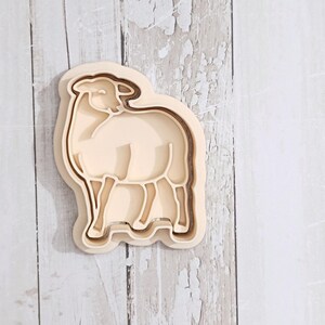 May include: A cream-coloured plastic cookie cutter shaped like a standing sheep. The cutter features a raised outer border and an inner recessed design that outlines the head, body, and four legs of the animal in a simple, clean silhouette.